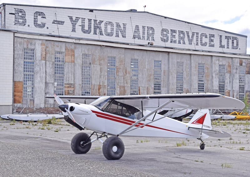 A World War II era hangar at Watson Lake, Yukon territory, provides the backdrop for a modern Carbon Cub EX enroute to Alaska. The Cub is owned by its builder, Daniel Lilja of Plains, Mont. (Photo by Bill Walker)